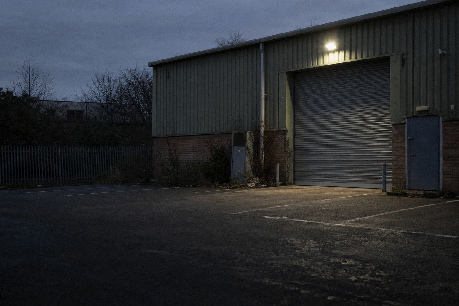 A closed industrial unit at dusk, the shutter down and one light on.