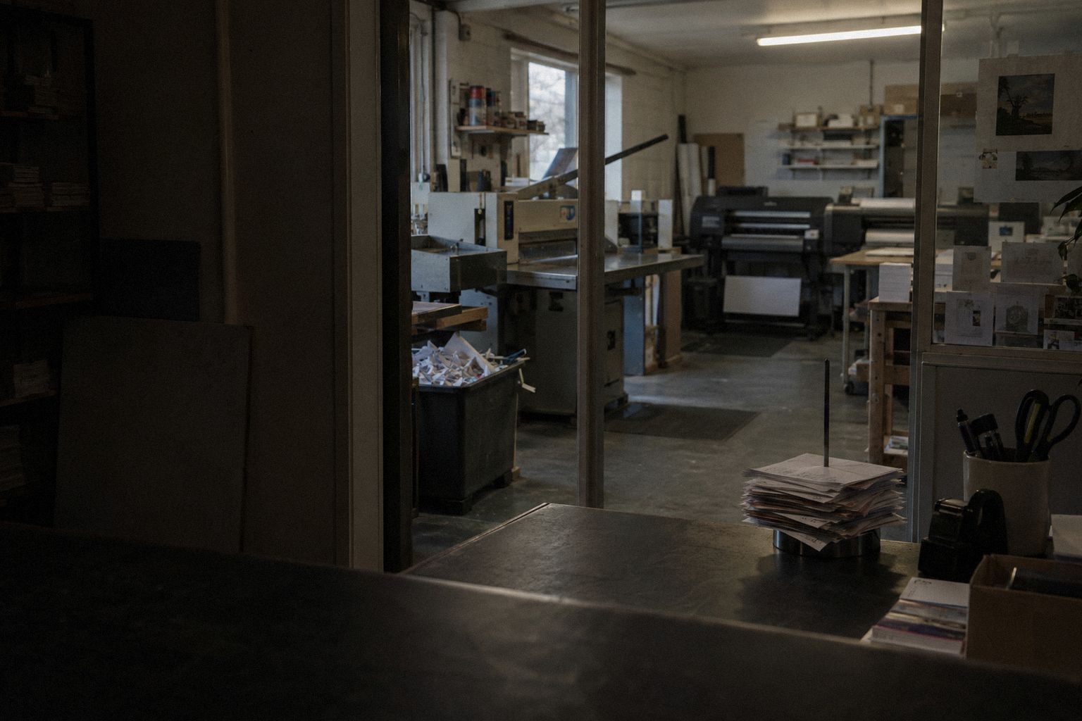 A print shop after closing, job tickets on a spike and the machines quiet behind glass.