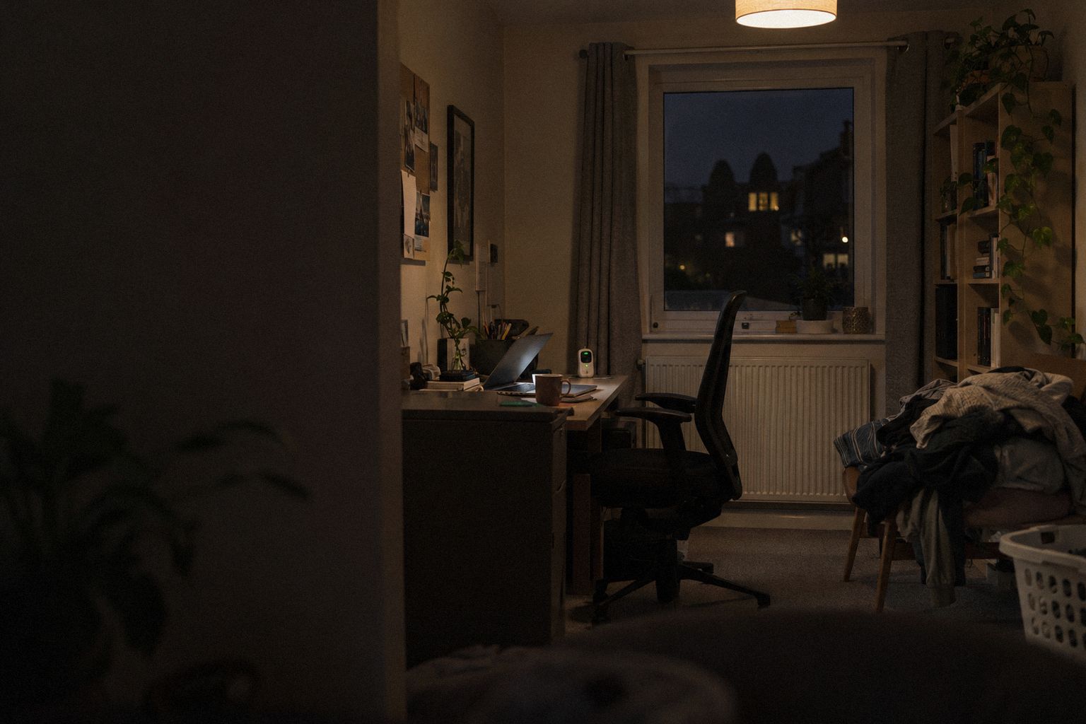 A box-room office at 9pm, a baby monitor beside a half-open laptop.