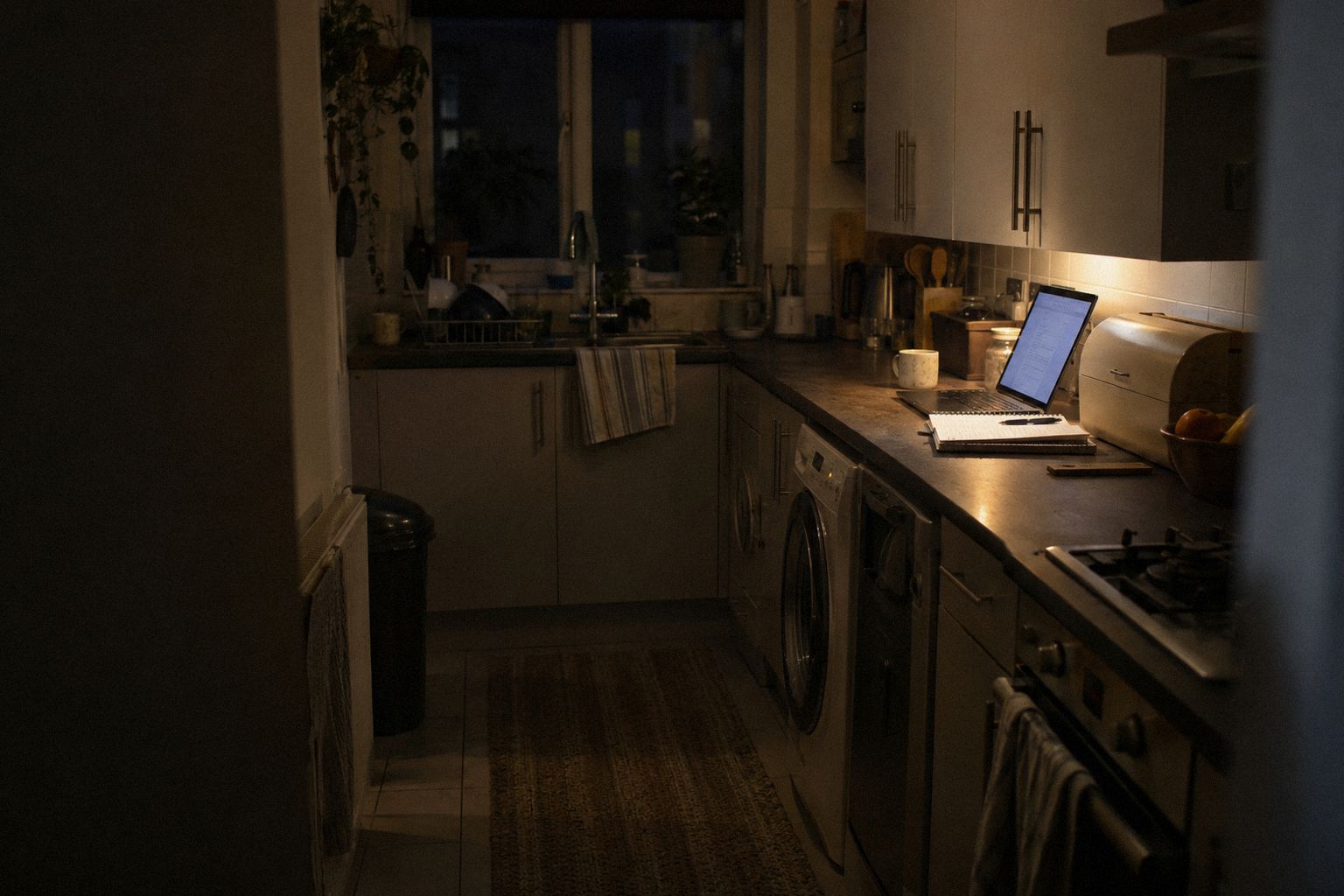 A galley kitchen late at night, a laptop on the worktop beside a notebook.