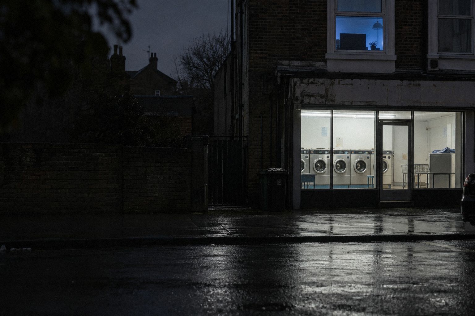 A launderette at night with a lit workroom window above it, rain on the street.
