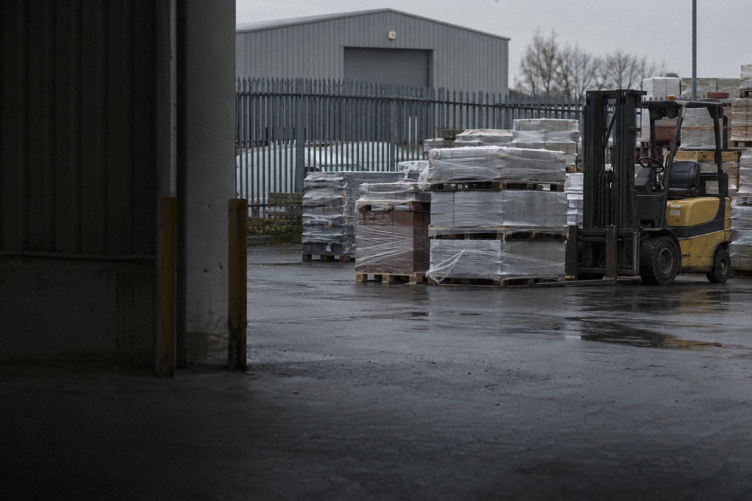Pallets of stock wrapped in plastic in a yard at first light, a forklift beside them.