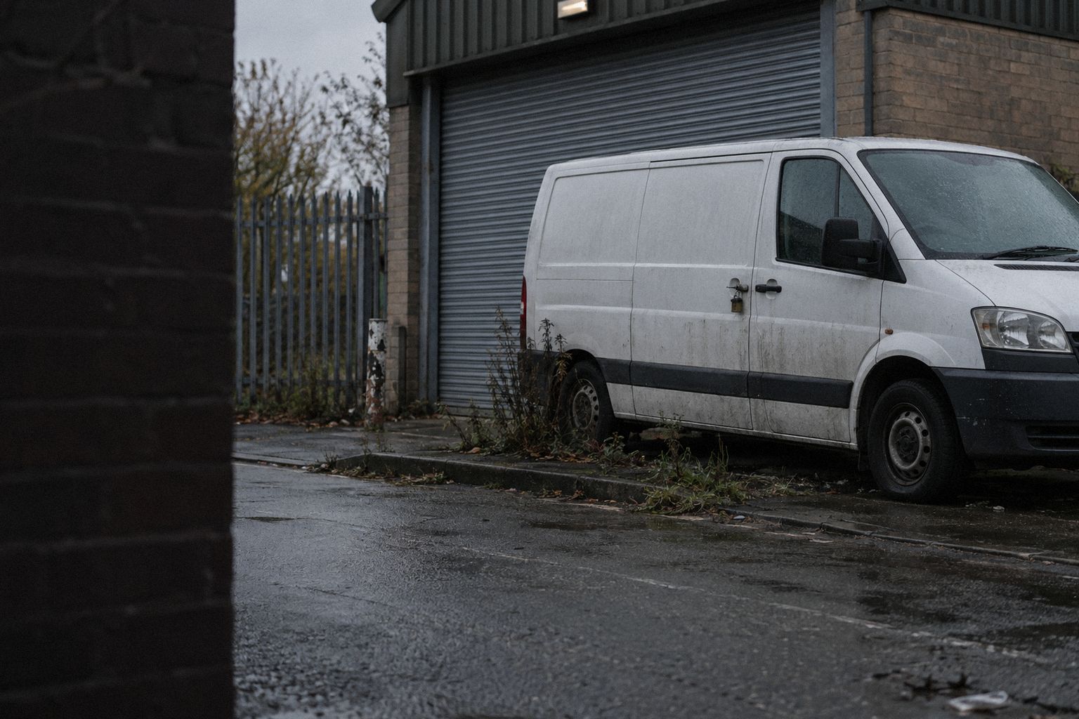 A van outside a closed workshop in the drizzle, the shutter down.