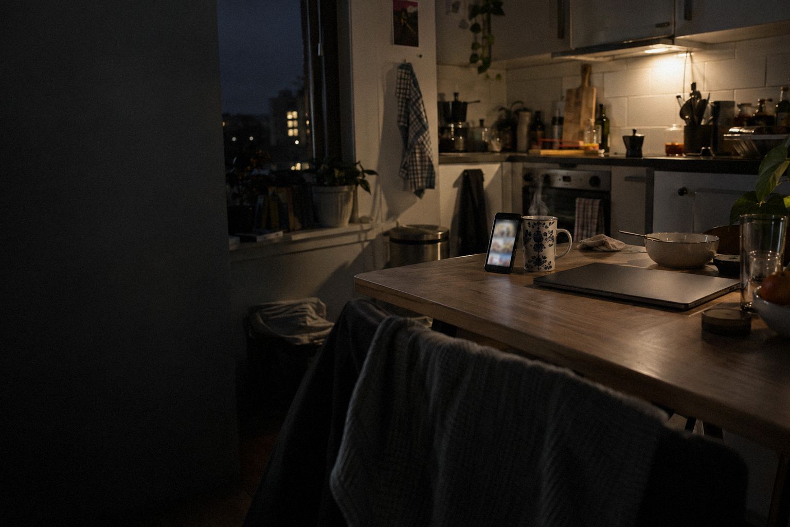 A studio kitchen at 9pm, a phone propped against a mug beside a closed laptop.