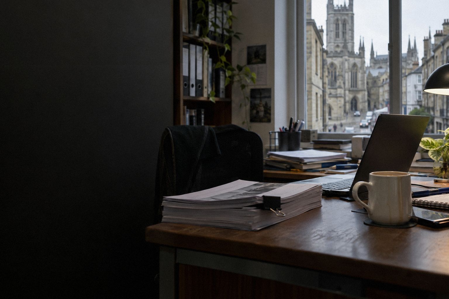 A thick printed tender pack on a desk at 4pm, a cold cup of tea beside it.