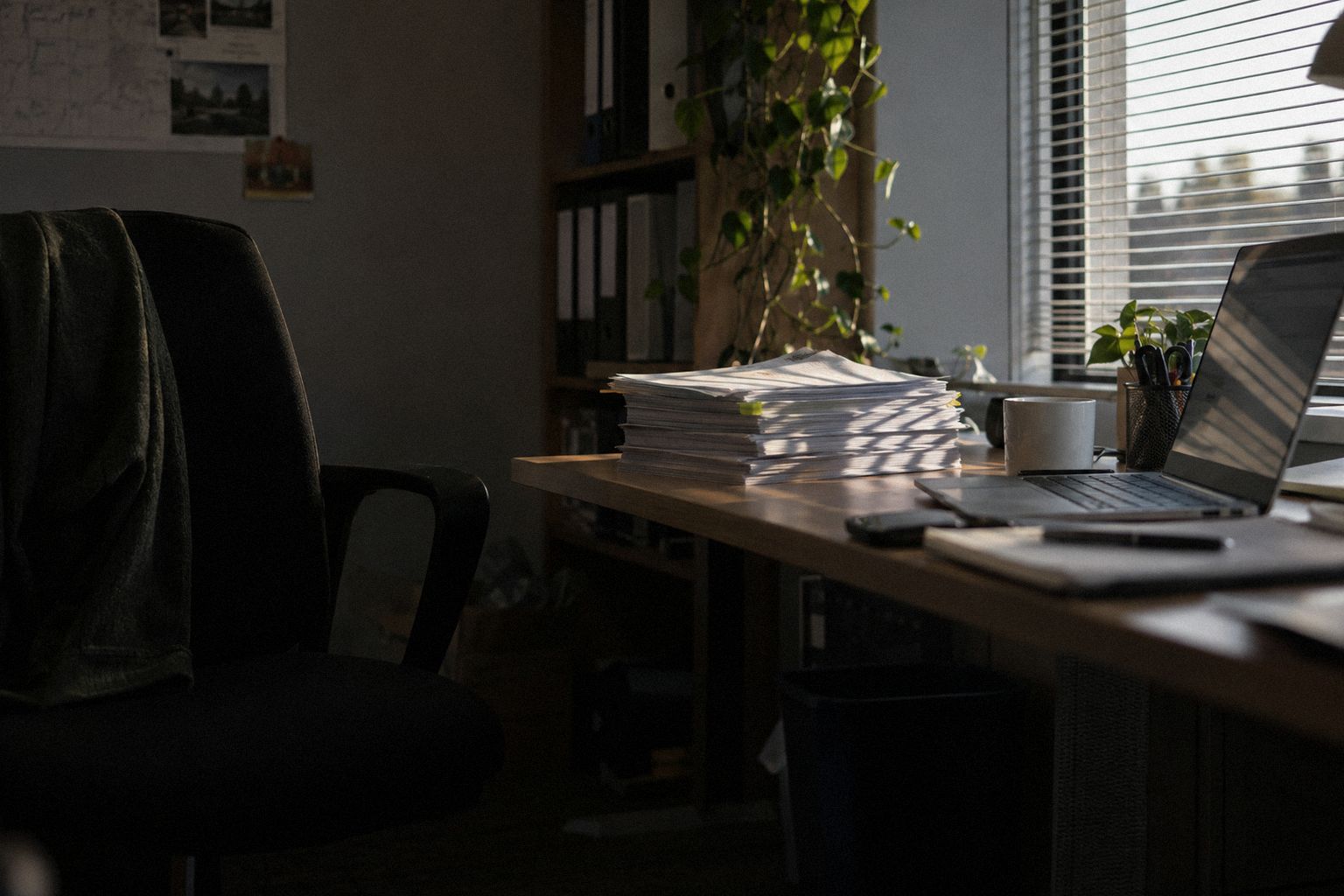 Low sun through blinds across a stack of papers on an office desk.
