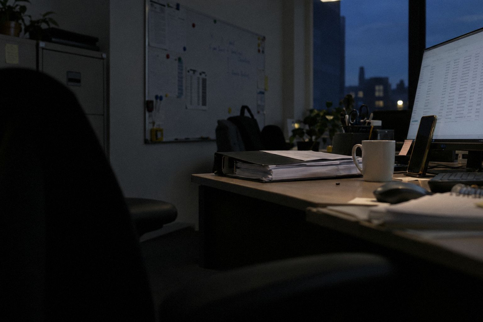 A back office at 6pm, a phone propped against the screen and an invoice folder.