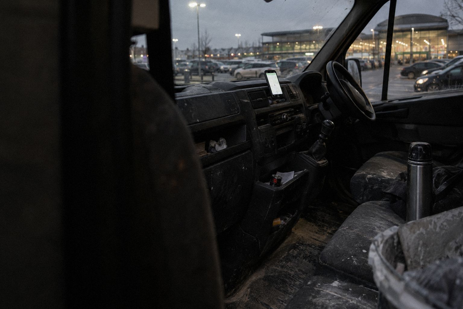 A plasterer's van in a supermarket car park at dusk, a phone glowing on the dashboard.