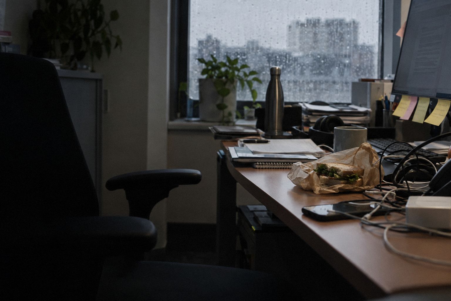 A cluttered desk at lunchtime, sticky notes on the screen and a sandwich in its wrapper.