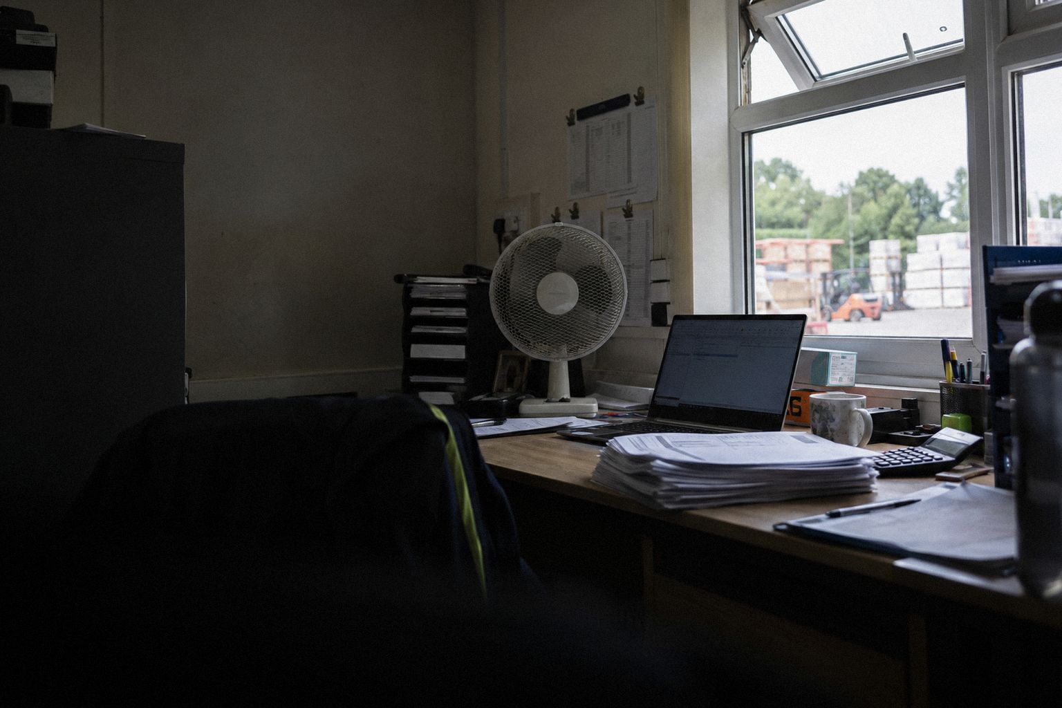 A back office in July, a desk fan turning beside a laptop and delivery notes.