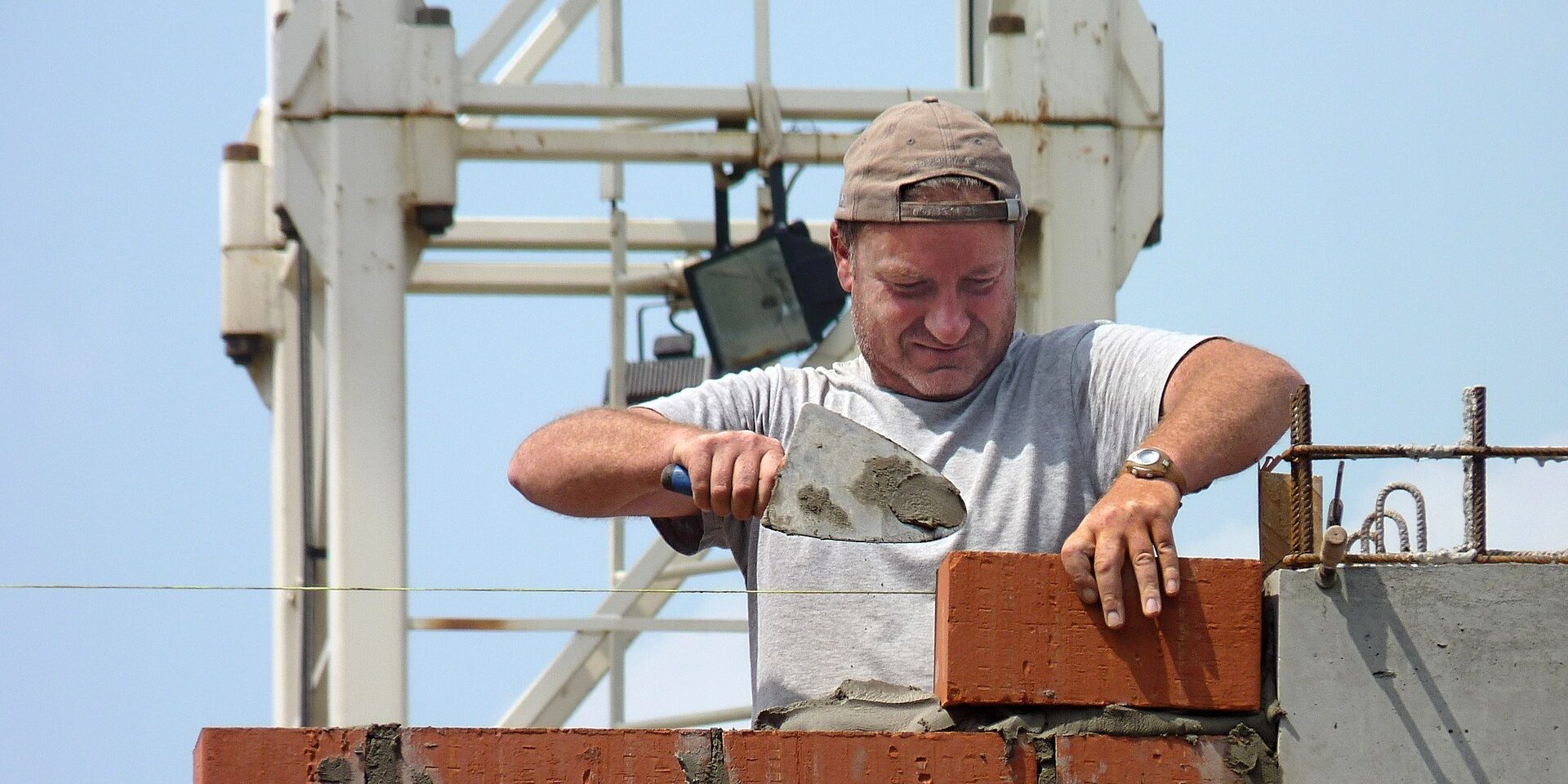 A bricklayer in a cap and a grey T-shirt lays a brick on a fresh course of mortar, a crane behind him.