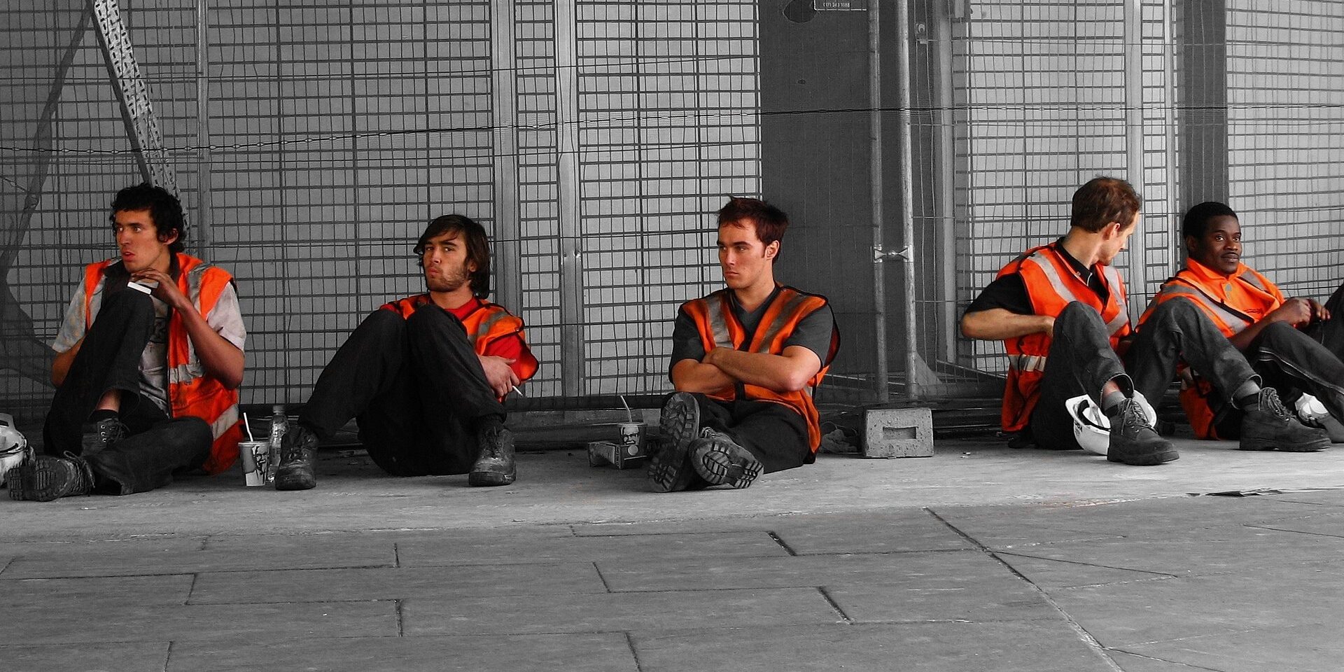 Five builders in orange hi-vis vests sit on the pavement against a site fence, on a break.