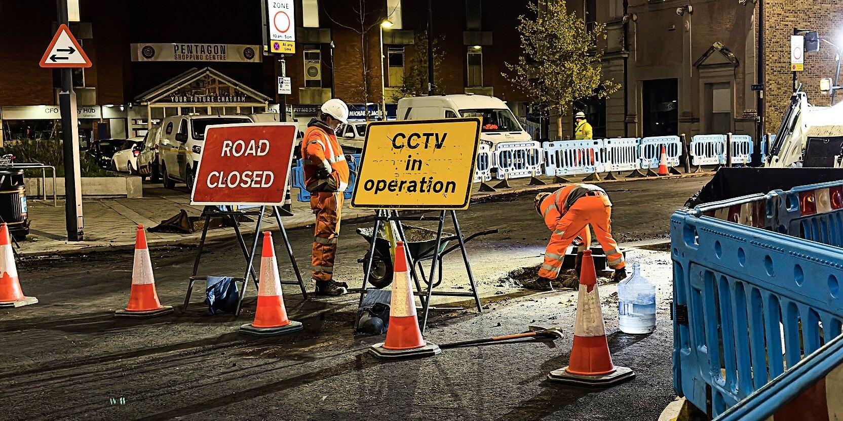 A road crew in orange hi-vis works at night behind barriers and a Road Closed sign, a digger lit up behind them.