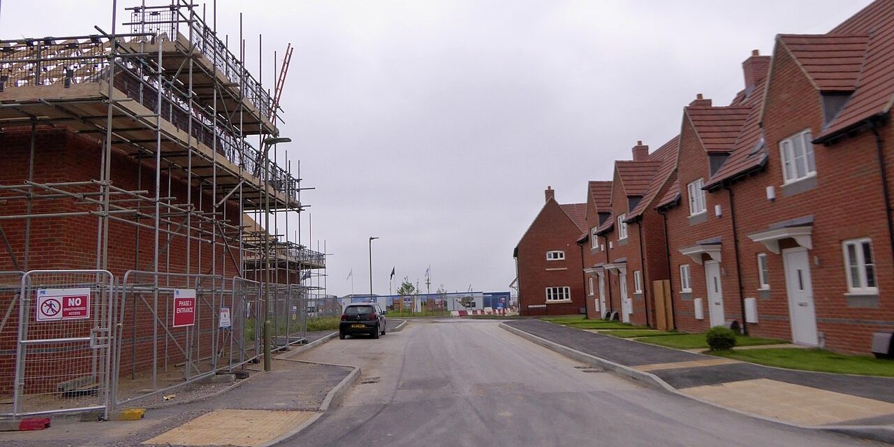 A new street of brick houses, the nearest still in scaffolding, behind site fencing.